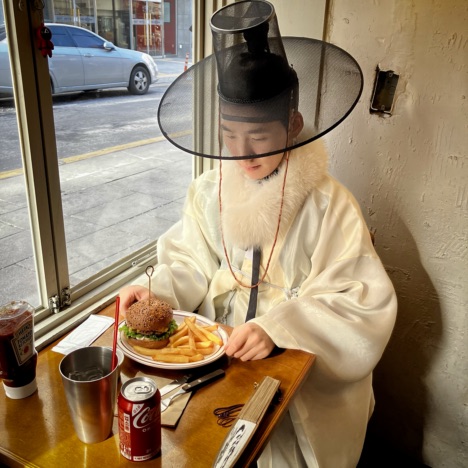 SuhJae wearing a yellow Hanbok (도포, traditional Korean clothing) while eating burgers.